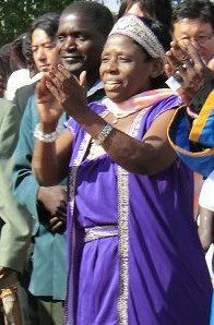 Reen Kachere. Eine afrikanische Frau in lila Kostüm mit einem Diadem klatscht vor einer Menschenansammlung in die Hände. 2013