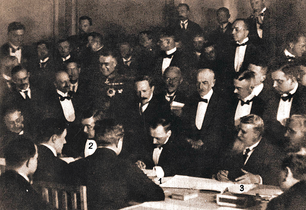 Three formally attired men at a conference table sign documents while 32 others look on.