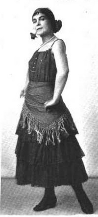 A white female dancer, standing with arms akimbo, wearing a dark, sleeveless dress, with a fringed scarf tied across her hips; she may have a flower in her mouth.