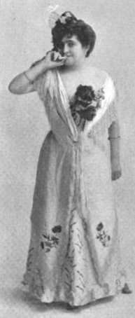 White woman standing in a light-colored gown with floral embellishments.