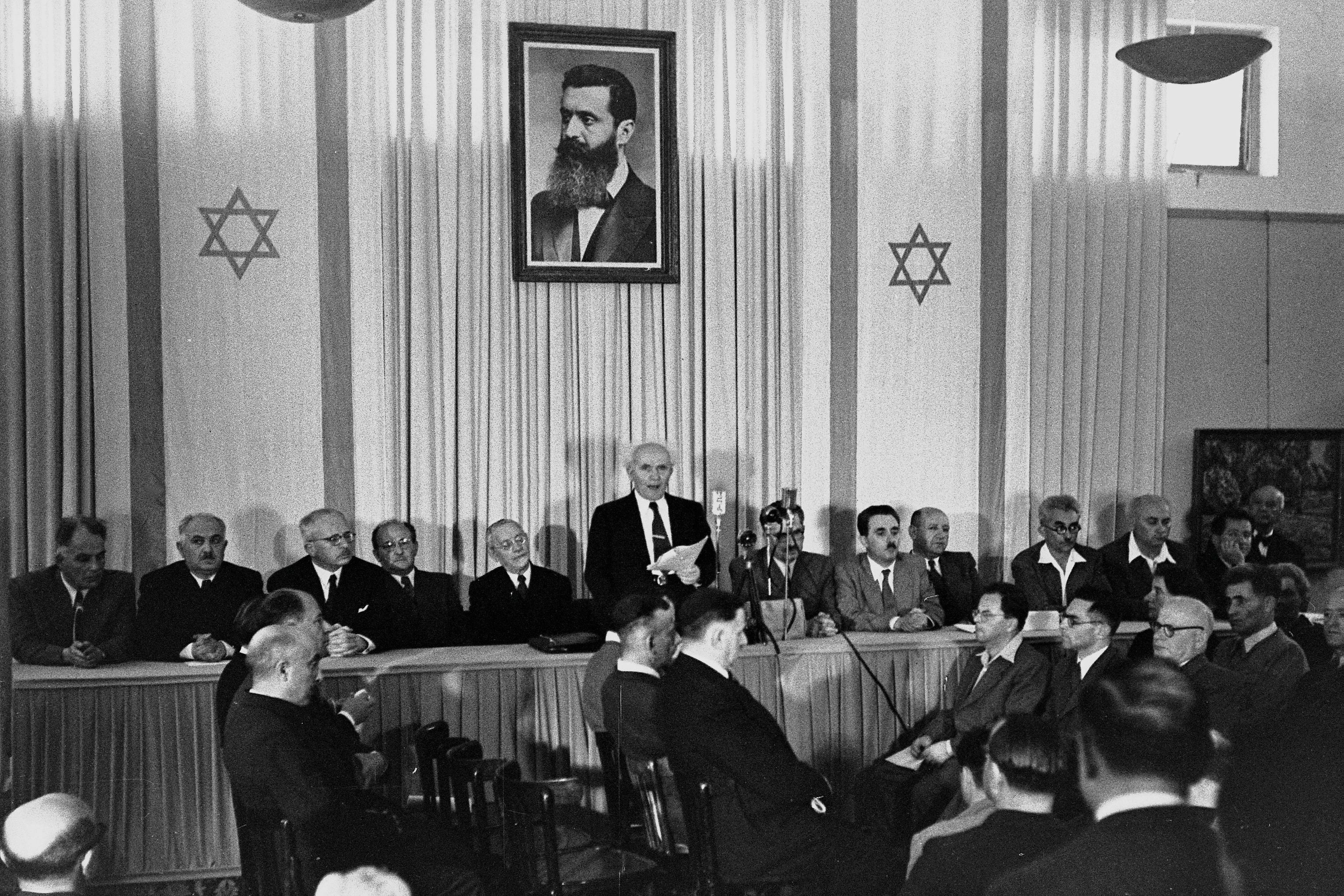 A single man, adorned on both sides by a dozen sitting men, reads a document to a small audience assembled before him. Behind him are two elongated flags bearing the Star of David and portrait of a bearded man in his forties.