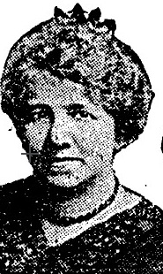 A newspaper photo of a middle-aged white woman, wearing a strand of dark beads and a v-necked top. Her hair is arranged in an updo with a crown-like ornament on the top.