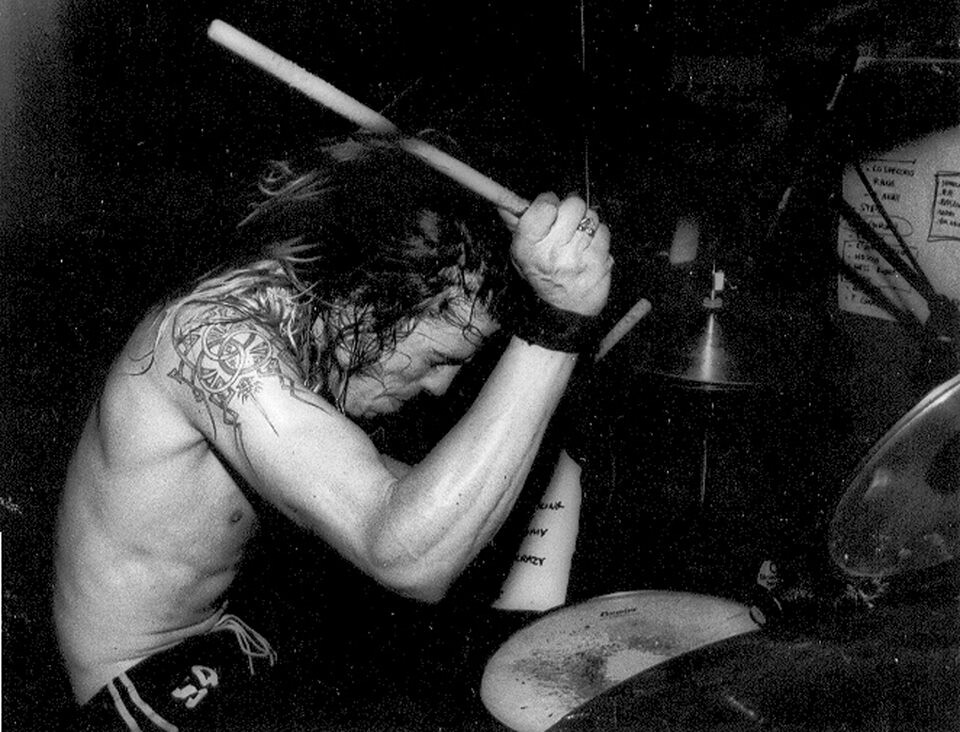 photo en noir et blanc d'un homme torse nu jouant violemment de la batterie.