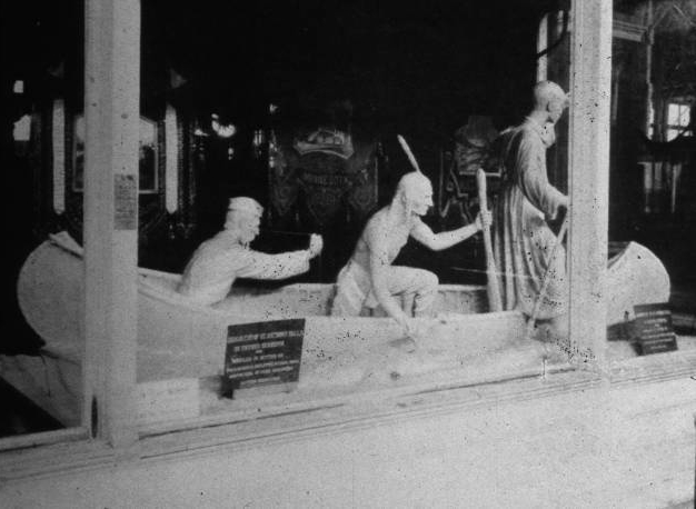 Photograph of an elaborate butter sculpture of a priest sitting in a boat with two guides exiting the boat, one of whom has clear indigenous American garb