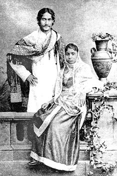 Black-and-white photograph of a finely dressed man and woman: the man, smiling, stands akimbo behind a settle with a shawl draped over his shoulders and in Bengali formal wear. The woman, seated on the settle, is in elaborate Indian dress and shawl; she leans against a carved table supporting a vase and flowing leaves.