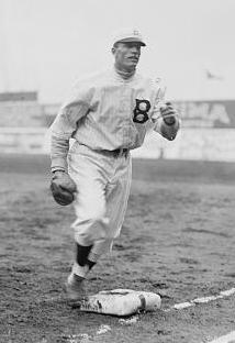 A man in a light baseball uniform with a dark "B" on the left chest running past first base with a fielding glove on his right hand