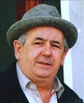 "Small coloured photograph of a man in his 60s wearing a blue shirt and a trilby hat"