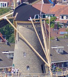 Photographie en couleurs de la partie supérieure d'un moulin à vent au milieu d'un paysage urbain, des personnages visibles sur sa galerie.
