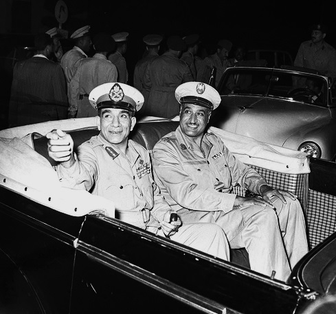 رجلان مبتسمان يرتديان الزي العسكري ويجلسان في سيارة مكشوفة. الرجل الأول على اليسار يشير بيده في لفتة. خلف السيارة رجال يرتدون الزي الرسمي يبتعدون عن السيارة