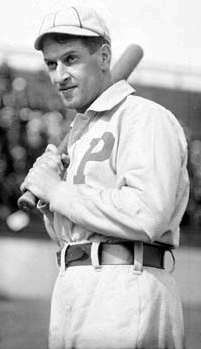 A man in an old-style white baseball uniform with a large block "P" over the left breast and crownless cap holds a baseball bat over his right shoulder.