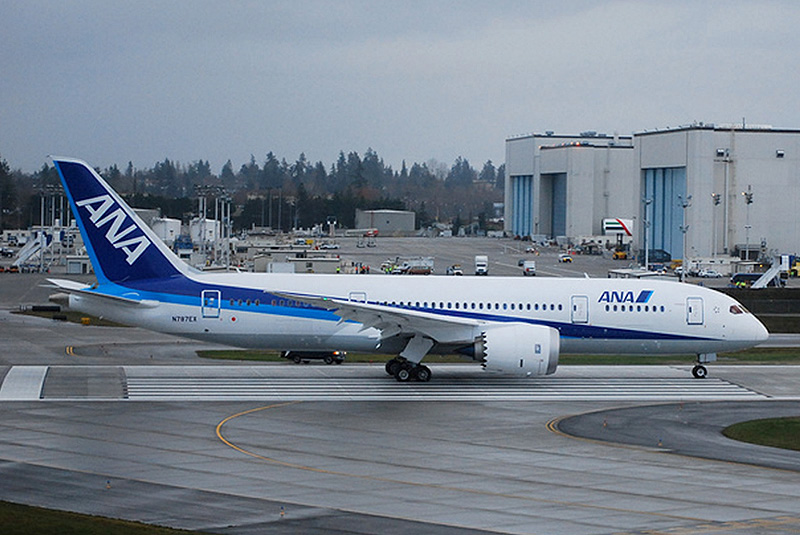 B787 in launch customer All Nippon Airways' blue and white livery. In the background are two assembly halls, with huge doors facing left. Vehicles are parked in front of the halls.