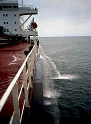 A cargo ship is pictured from the front, with water being pumped out of the sides of the ship, near the top railing.