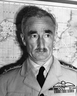 Portrait de tête et d'épaules d'un homme moustachu en uniforme militaire clair avec des ailes de pilote sur la poche gauche.