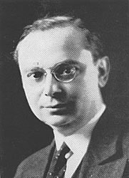 Portrait d'un homme rasé de près avec des cheveux partant sur le côté gauche, portant des lunettes, un costume et une cravate.