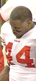 An American football player looks down. He is not wearing his helmet. He is wearing a white jersey with the number 44 across the chest.