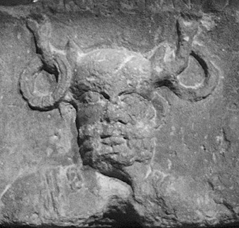 a rough and old carving shows a man's face with horns, each of which carries a large hoop or ring