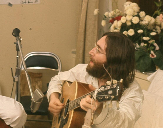 A bearded, bespectacled man in his late twenties, with long dark brown hair and wearing a loose-fitting pajama shirt, sings and plays an acoustic guitar. White flowers are visible behind and to the right of him.
