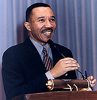 An African-American male with a mustache wearing a black suit with a light blue shirt and a red and white diagonally striped tie smiles as he stands behind a podium and adjusts a microphone with his right hand.