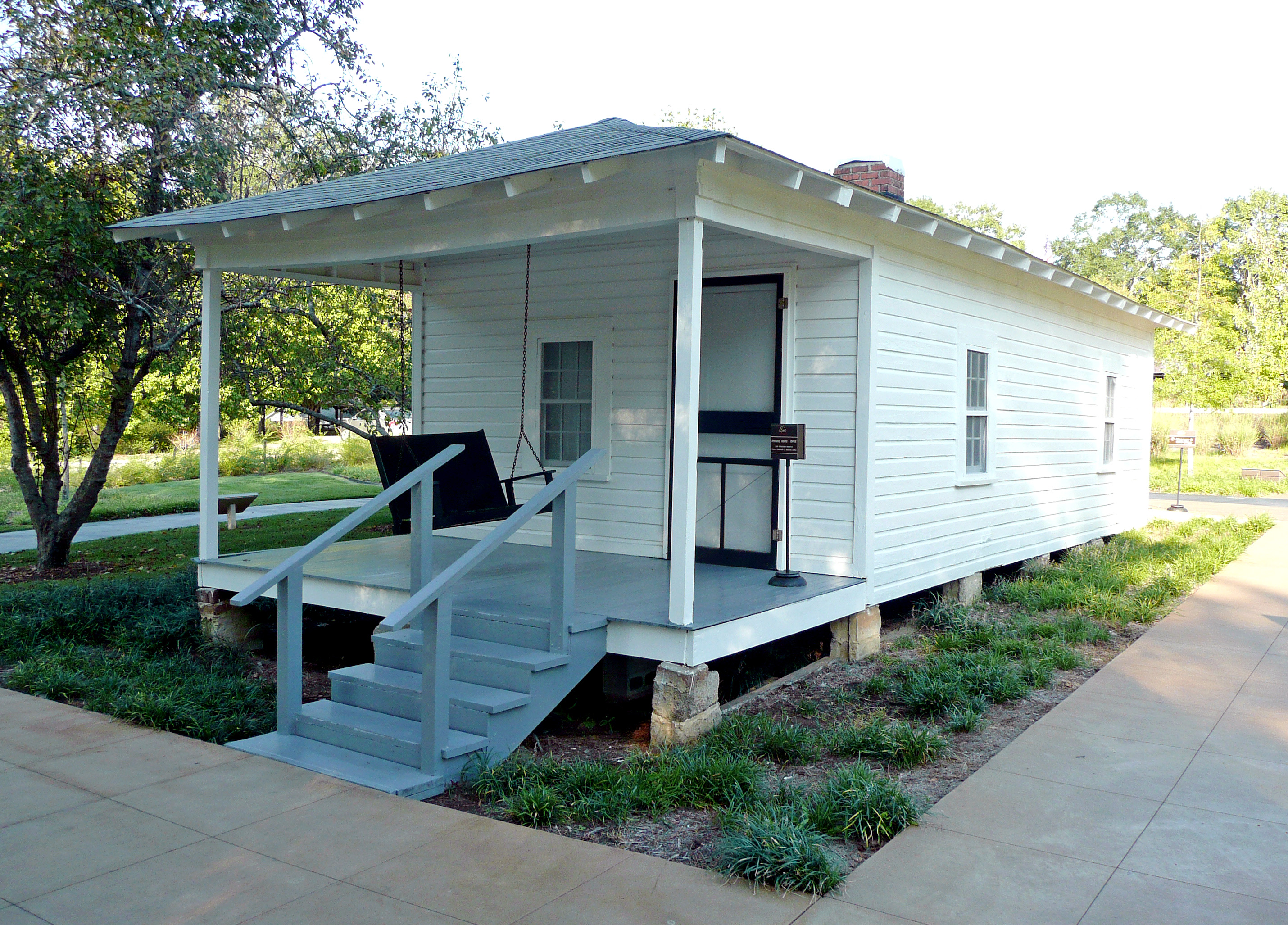 Present-day photograph of a whitewashed house, about 15 feet wide. Four bannistered steps in the foreground lead up to a roofed porch that holds a swing wide enough for two. The front of the house has a door and a single paned window. The visible side of the house, about 30 feet long, has two paned windows.