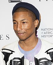 A color photograph of Pharrell Williams, who is standing in front of a step and repeat.