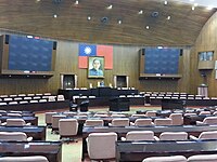 Photo of a chamber with rows of seats, with a podium at the front and a large portrait and flag overhead