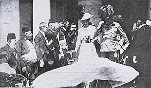 Černobílá fotografie manželů d'Este sestupujících po schodech; arcivévoda v generálské uniformě napravo, vévodkyně v bílých šatech se širokým kloboukem nalevo. Kolem špalír přihlížejících.