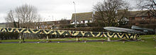 A grey rocket on a camouflaged ramp in front of a hedge. The tops of houses and street lights are visible behind the hedge.