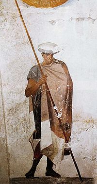 Fresco of an ancient Macedonian soldier with a grounded spear. He wears three items of clothing, which, in combination, are considered typical of Macedonians: The kausia cap, the Macedonian type of chlamys (cloak) and krepides (boots), from the tomb of Agios Athanasios, Thessaloniki, Greece. Fresco of a Macedonian soldier, from the Tomb of Agios Athanasios, 4th century BC.jpg