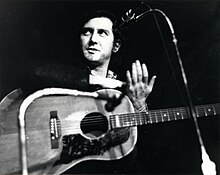 Photo en noir et blanc d'un homme muni d'une guitare derrière un micro à pied