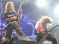 A color photograph of two members of the group Children of Bodom standing on a stage with guitars, drums are visible in the background. Both electric guitarists have "flying V" style guitars and they have long hair.