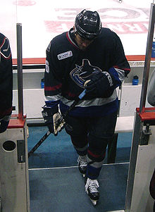 An ice hockey player dressed in a black jersey. He is walking on the players' bench and looking downwards.