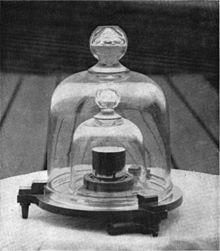 Černobílá fotografie, kde můžeme vidět prototyp kilogramu postaveném na stolku.