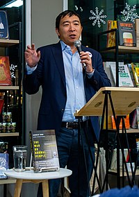 Yang is holding a microphone while gesturing and making a speech. His book, The War on Normal People, is displayed on a table in front of him.