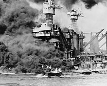 Dark smoke billows from the ship after the Pearl Harbor attack while a man is rescued from the water by sailors in a small boat