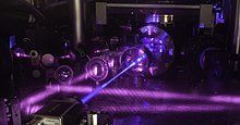 JILA's 2017 three-dimensional (3-D) quantum gas atomic clock consists of a grid of light formed by three pairs of laser beams. A stack of two tables is used to configure optical components around a vacuum chamber. Shown here is the upper table, where lenses and other optics are mounted. A blue laser beam excites a cube-shaped cloud of strontium atoms located behind the round window in the middle of the table. Strontium atoms fluoresce strongly when excited with blue light. 17jila003 3d strontium atomic clock.jpg