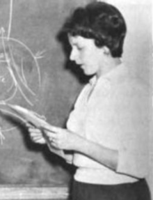 A young white woman with short dark hair, standing in front of a chalk board, holding papers; she is wearing a white blouse