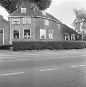 Photographie en noir et blanc d'une maison couronnée d'une galerie et longée par une route.