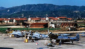 Three single-seat, piston-engined fighters under maintenance, with red-roofed building and a mountain range in the background