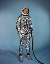 Armstrong standing up, wearing an early space suit. It is highly reflective silver in appearance. He is wearing the helmet, which is white, with the visor raised. A thick dark hose is connected to one of the two ports on the front abdomen of the suit.