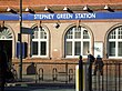 A red-bricked building with a rectangular, dark blue sign reading "STEPNEY GREEN STATION" in white letters and a man walking in front