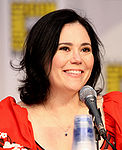 A woman with black hair, tied back, smiling, and sitting behind a microphone