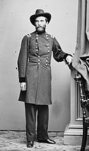 Black and white photo shows a bearded man in a broad-brimmed hat standing full-length. He wears a dark military uniform with two rows of buttons. The coat reaches down to the knees.