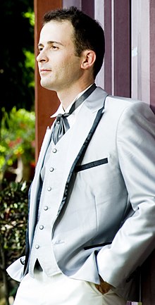 Темноволосый мужчина во фраке и камербанде с темным галстуком-бабочкой, обе руки в карманах светлых брюк.