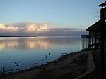 A view of a large bed of water near sunset. Onshore to the right is a small shack made of a thin material elevated with wooden poles.