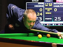 Photograph of John Higgins wi a cue in haund, ready tae strike the cue ball.