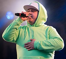 Man wearing a baseball cap and green hoodie, performing onstage with a microphone in hand