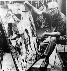 Vaclav Vytlacil seated in a chair holding a medium sized abstract painting to look at