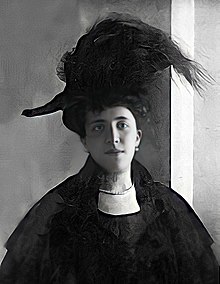 Photographie noir et blanc d'une femme, vêtue de noir et portant un grand chapeau avec des plumes.