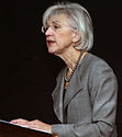 A colour photograph of a woman with white hair facing the left and wearing glasses, a grey dress jacket, and a silver-beaded necklace