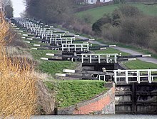 A series of approximately 20 black lock gates with white ends to the paddle arms and wooden railings, each slightly higher than the one below. On the right is a path and on both sides grass and vegetation.